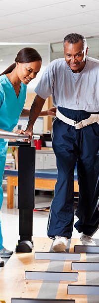 Physical therapist (40s) helping patient (60s) through gait training. Physical therapist (40s) helping patient (60s) through gait training.